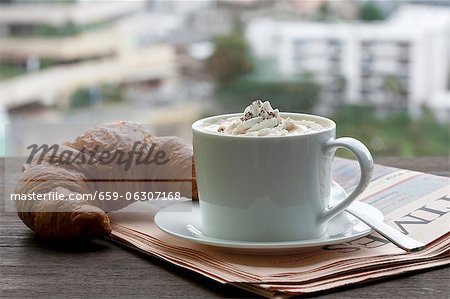 A cappuccino with a croissant on newspaper in front of a window