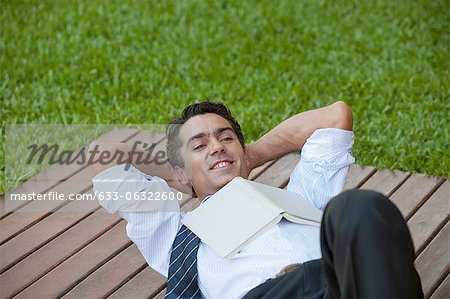 Mid-adult man lying outdoors with hands behind head and book on chest