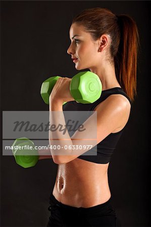 Photo of a slim female lifting dumbbells doing bicep curls over black background.