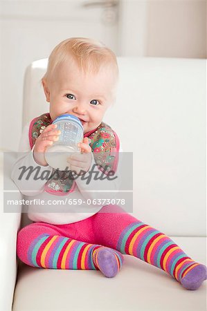 A baby sitting on a sofa with a bottle of milk