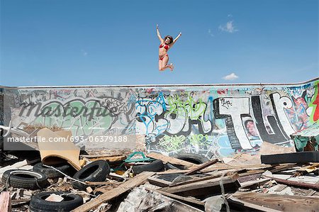 Young woman in bikini jumping over graffiti wall with garbage in foreground