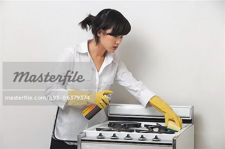 Young housemaid cleaning stove against gray background