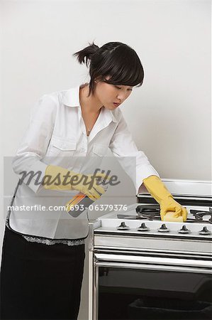 Young maidservant cleaning stove against gray background