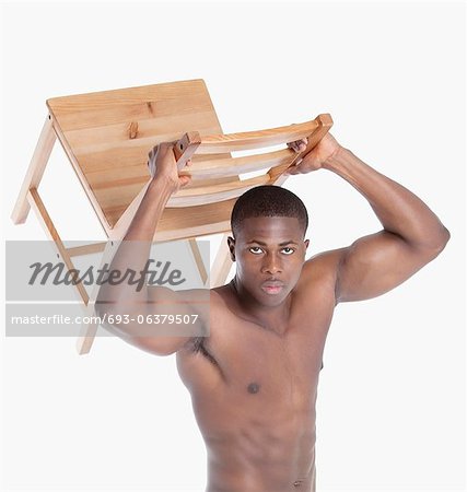 Portrait of a shirtless young male athlete lifting wooden chair over gray background