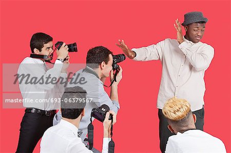 Young male celebrity shielding face from photographers over red background