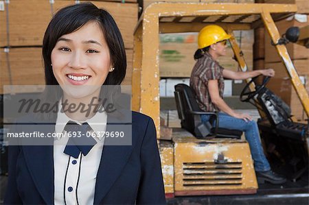 Portrait of woman smiling while female industrial worker driving forklift truck in background