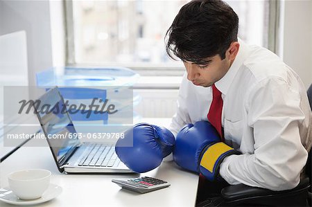 Young Indian businessman wearing boxing gloves while using calculator at office desk
