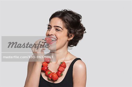 Glückliche junge Frau Erdbeere Halskette tragen, wie sie ein Stück über den grauen Hintergrund isst