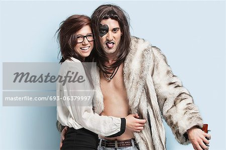 Young man in fur coat sticking out tongue while standing with happy woman against light blue background