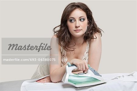 Young woman looking away while ironing shirt over gray background