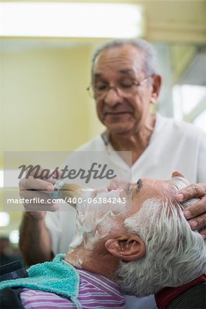 Elderly barber with shave brush applying cream to client in old style shop