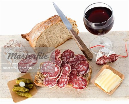 Sliced French saucisson on bread with knife, wine and pickels, in front of white background