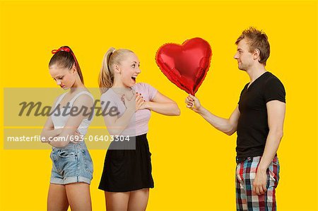 Young man gifting heart shaped balloon to surprised woman with friend feeling left out standing behind