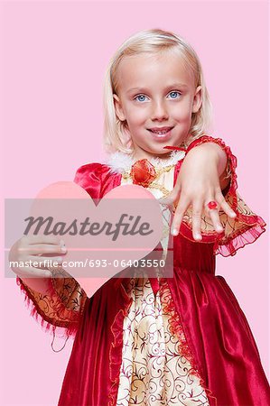 Portrait d'une jeune fille heureuse habillé en costume de princesse holding coeur en papier, car elle affiche anneau sur fond rose