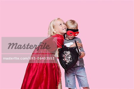 Happy young girl in princess costume hugging boy pretending to be her hero over pink background