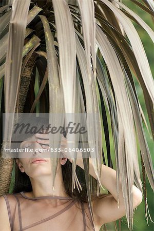 Young woman behind tropical foliage, hand covering one eye