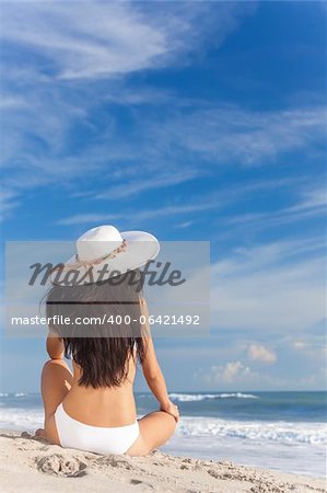 Ein sexy junge Brünette Frau oder Mädchen trägt einen weißen Bikini und Sonne Hut sitzt auf einem einsamen tropischen Strand mit einem blauen Himmel