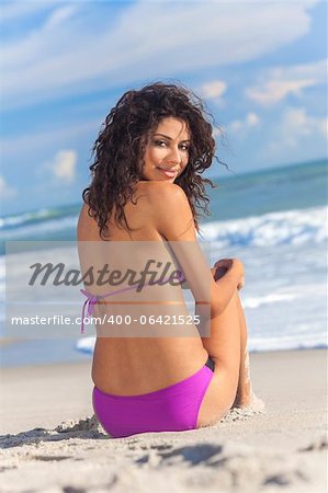 A sexy young brunette woman or girl wearing a purple bikini and sitting on a deserted tropical beach with a blue sky