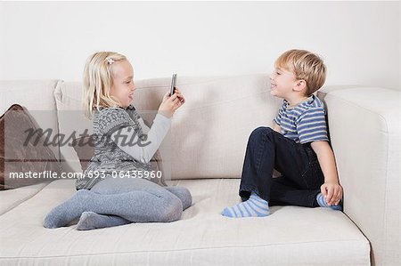 Young girl photographing brother through cell phone on sofa