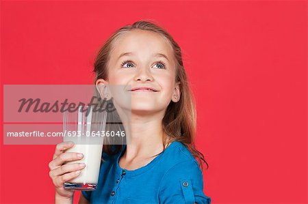 Young girl holding glass of milk while looking up on red background
