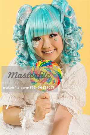 Portrait of smiling young woman dressed as a doll holding lollipop over yellow background