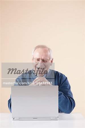 Senior Man Sitting at Table looking at Laptop Computer Laughing, Studio Shot on Beige Background