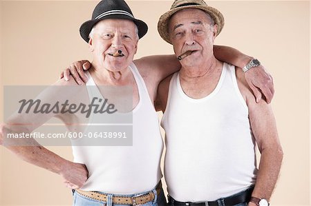 Portrait of Two Senior Man wearing Undershirts and Hats while Smoking Cigars with Arms around Shoulders, Studio Shot on Beige Background