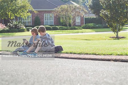 Two Boys Sitting on Neighbourhood Curb with Handheld Electronics