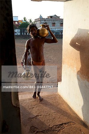 Monk, follower of Mahima Dharma sect, wearing orange loincloth carrying brass pots of water into the temple at sunset, Joranda, Orissa, India, Asia