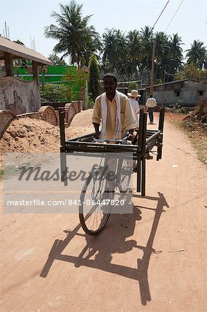 Man cycling through village street with two metal bed frames balanced on his bicycle, Hirapur, Orissa, India, Asia