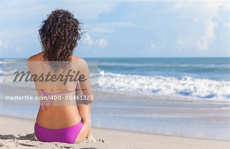 Rear view of a sexy young brunette woman or girl wearing a bikini sitting on a deserted tropical beach with a blue sky