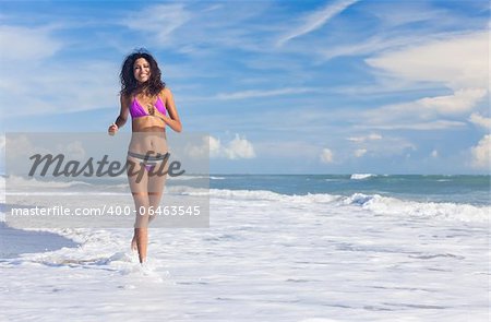 A sexy young brunette woman or girl wearing a bikini running through the surf on a deserted tropical beach with a blue sky