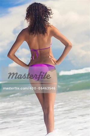 A sexy young brunette woman or girl wearing a bikini standing in the sea surf on a deserted tropical beach with a blue sky