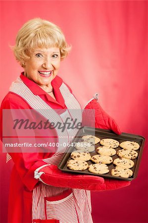 Sweet homemaker grandma holding a tray of fresh baked chocolate chip cookies.  Photographed in front of red background.