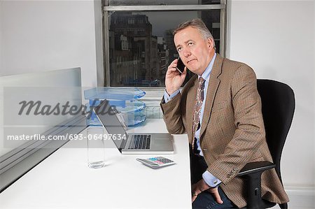 Middle-aged businessman using cell phone in front of laptop at desk in office
