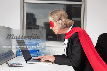 Side view of senior businesswoman in superhero costume using laptop at office desk