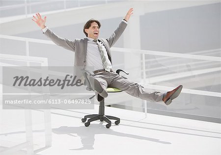 Carefree businessman sliding down walkway on office chair with wheels Carefree businessman sliding down walkway on office chair with wheels