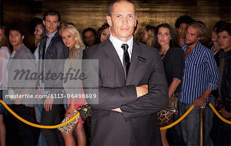 Portrait of serious bouncer with arms crossed in front of queue at nightclub