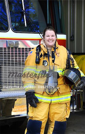 Fire Woman stands in front of fire truck wearing bunking uniform and holding helmet.  She is laughing as she is fully suited-up and ready for any fire emergency.