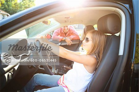 Young girl sitting in driver's seat of car, pretending to be old enough to drive as her smiling father watches on on a sunny summer evening in Portland, Oregon, USA