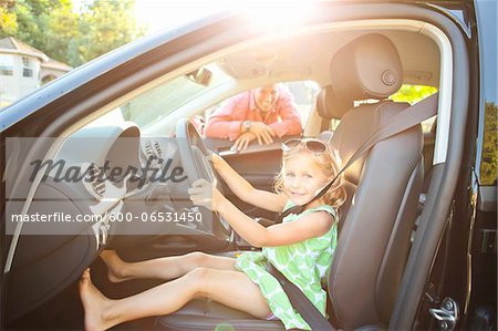 Little girl sitting in driver's seat of car wearing seatbelt, pretending to be old enough to drive and showing she knows the importance of a seat belt as her smiling father watches on on a sunny summer evening in Portland, Oregon, USA