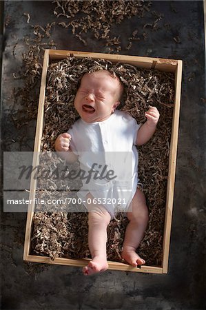 crying newborn baby girl in a white undershirt onesie in a wooden shipping box with cardboard packing paper