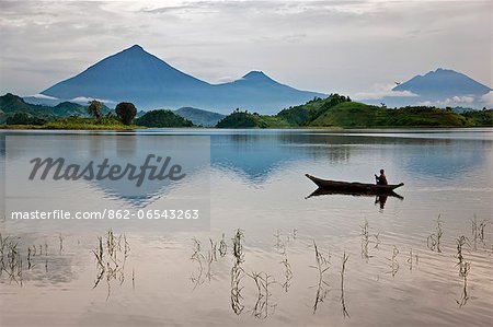 A woman paddles a dugout canoe across Lake Mutanda with its stunning backdrop of the Virunga Volcanoes, Uganda, Africa