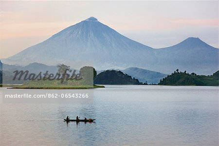 A man and woman paddle a dugout canoe across Lake Mutanda at sunrise. This lake with its many small islands has a stunning backdrop of the Virunga Volcanoes, Uganda, Africa