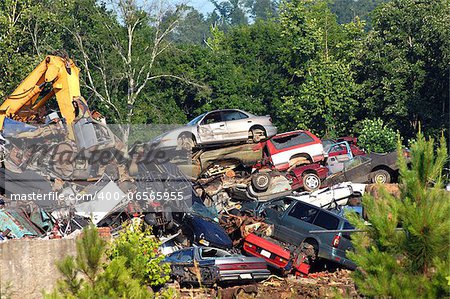 A pile of wrecked cars and auto bodies are crushed and stacked for spare parts or metal salvage.  Trees and weeds surround pile.