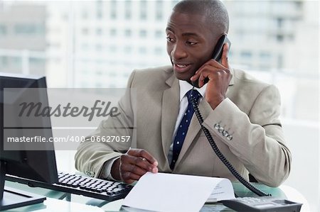 Smiling entrepreneur making a phone call while looking at his computer in his office