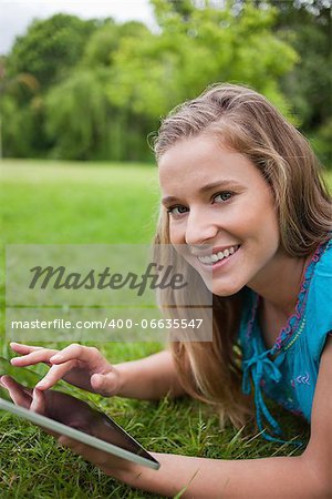 Smiling teenager lying down on the grass in a parkland while touching her tablet pc