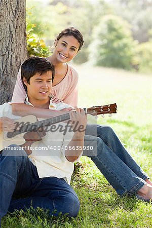 Woman sitting behind her friend and resting her arm on his chest while he plays a guitar under a tree