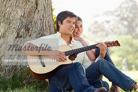 Man and his friend look into the distance as they listen to him playing the guitar while sitting against the trunk of a tree