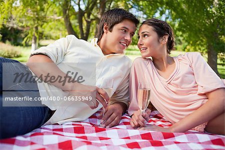 Two friends smiling as they look at each other while they lie down on a blanket and holding glasses of champagne